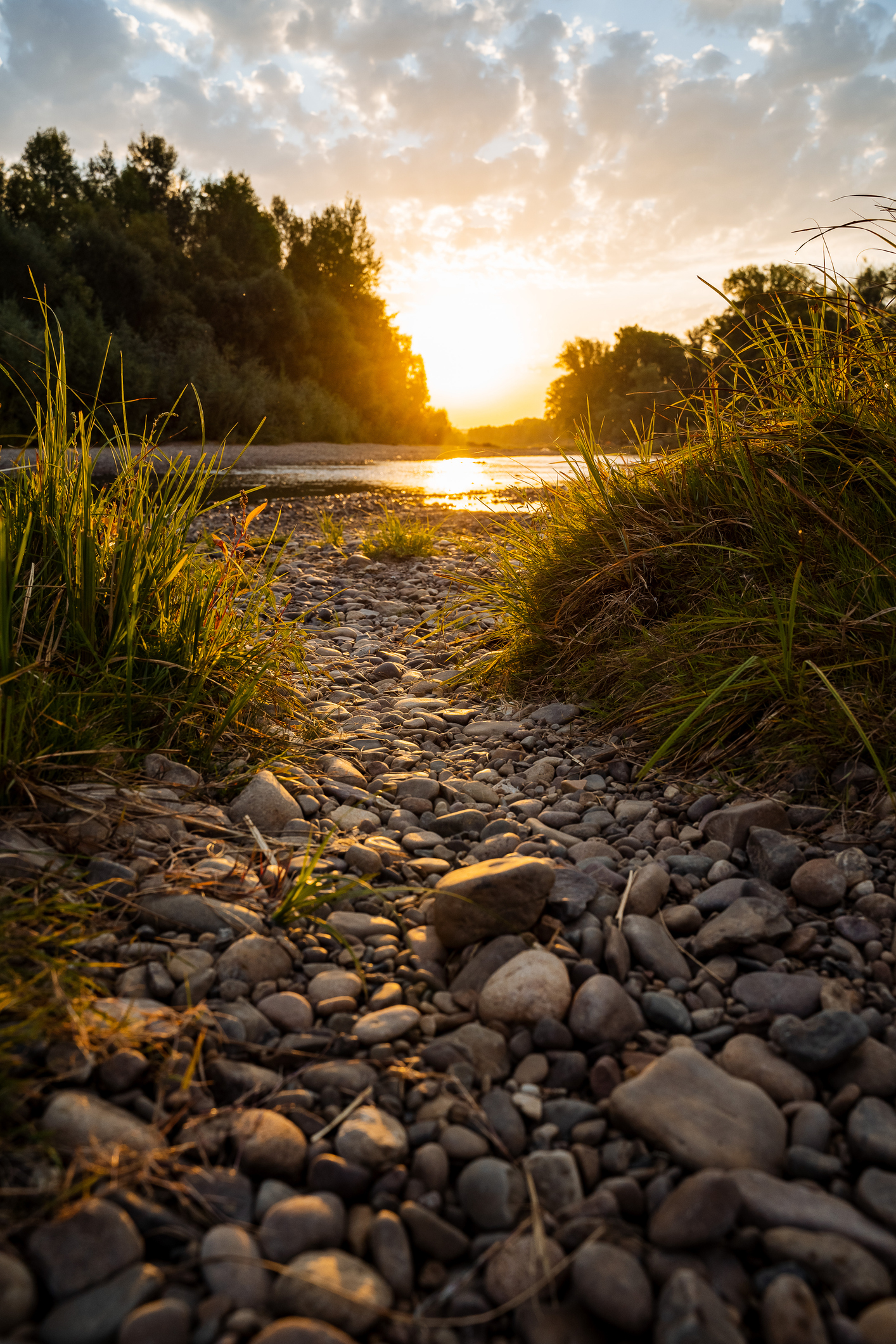 Incredibly beautiful sunset by the river. Rocky shore, trail leading to water. In the water surface reflected the sun’s rays. Outdoor activities in summer, hiking to the river